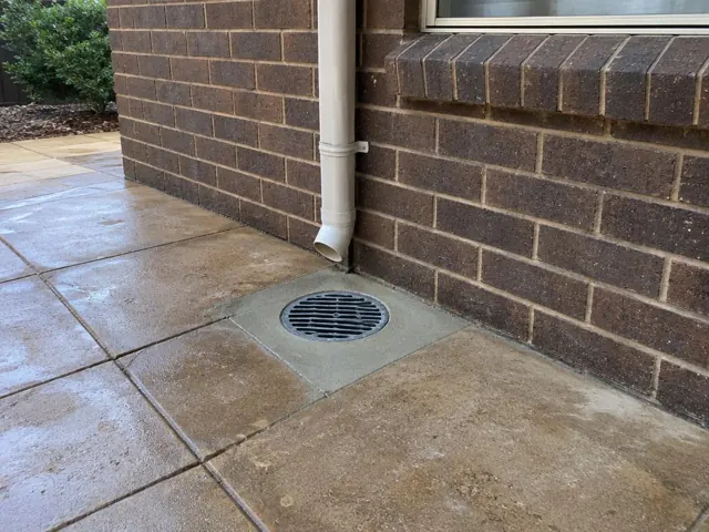 Outdoor drain grate beside brick wall and downpipe on dry paved patio in North Rocks